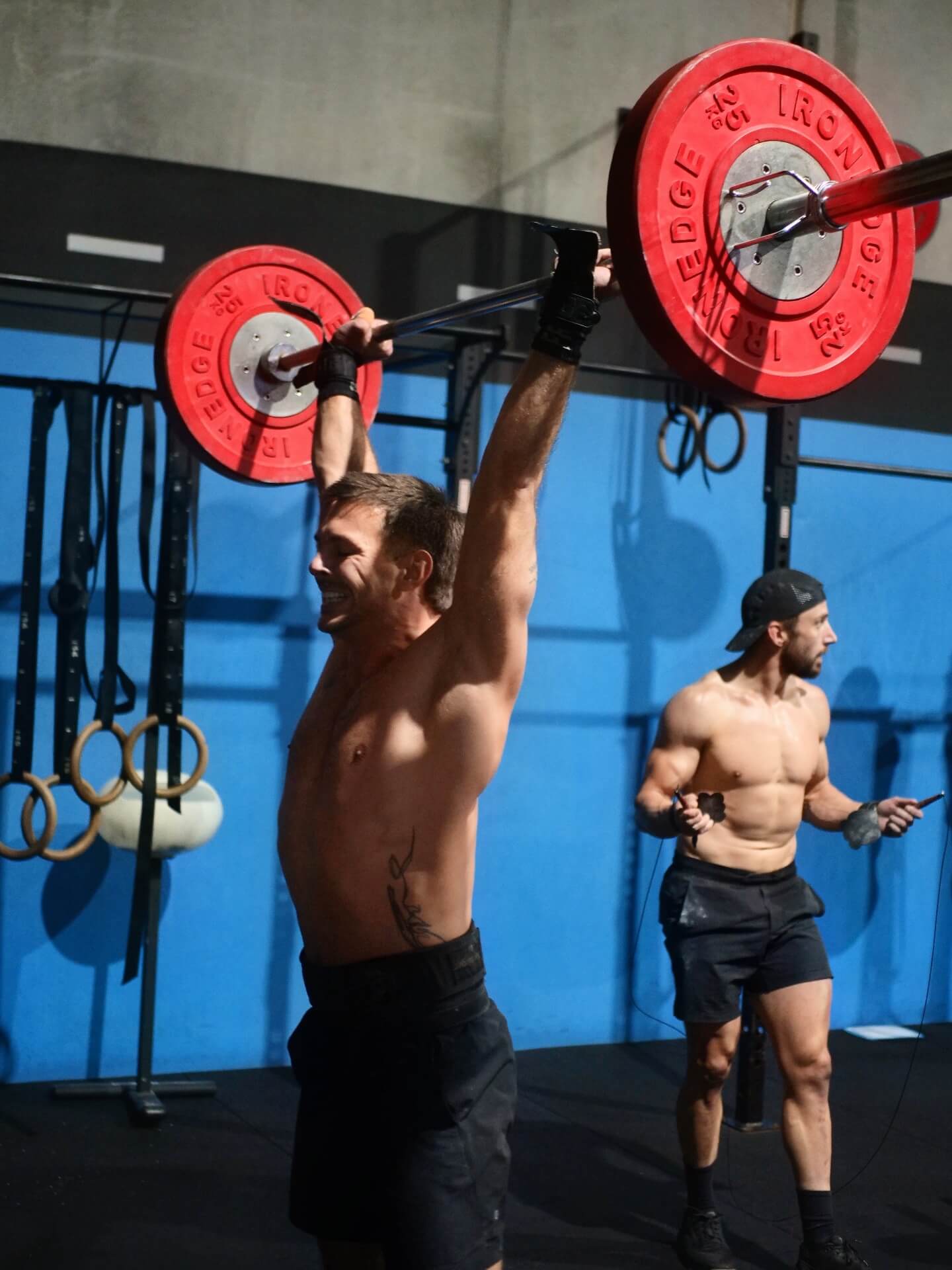 A member locking out a barbell overhead while another skips rope